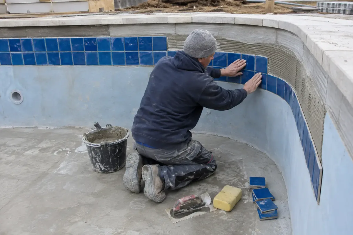 Specialist fitting blue ceramic tiles around the curved wall of an empty swimming pool, with pool floor and tile adhesive visible.